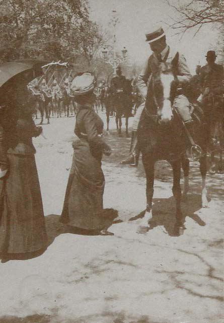 (Mujer conversando con un hombre a caballo. Desfile de militares detrás)