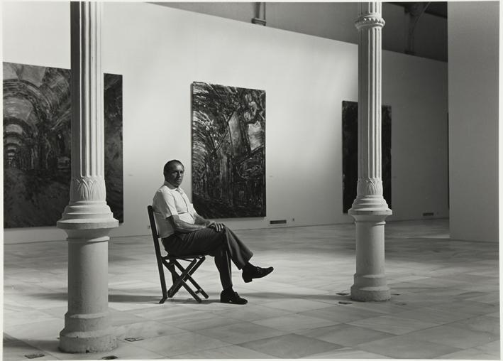 Retrato de hombre mayor sentado en una silla en una sala expositiva